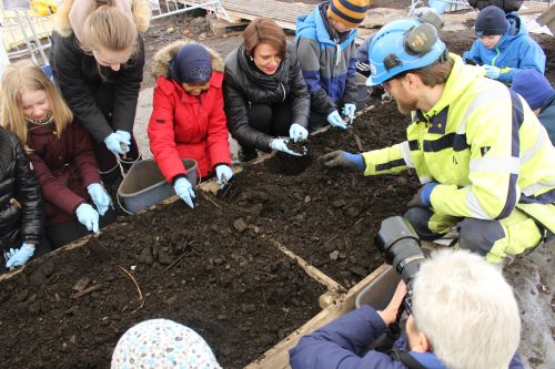Children wrapped up from cold weather excavating in a wooden trough supervised by an archaeologist wearing blue helmet and hi-vis jacket