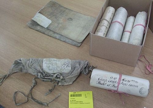 Parchment rolls in a box with one more on a table along with a grey coloured storage pouch and cataloguing label
