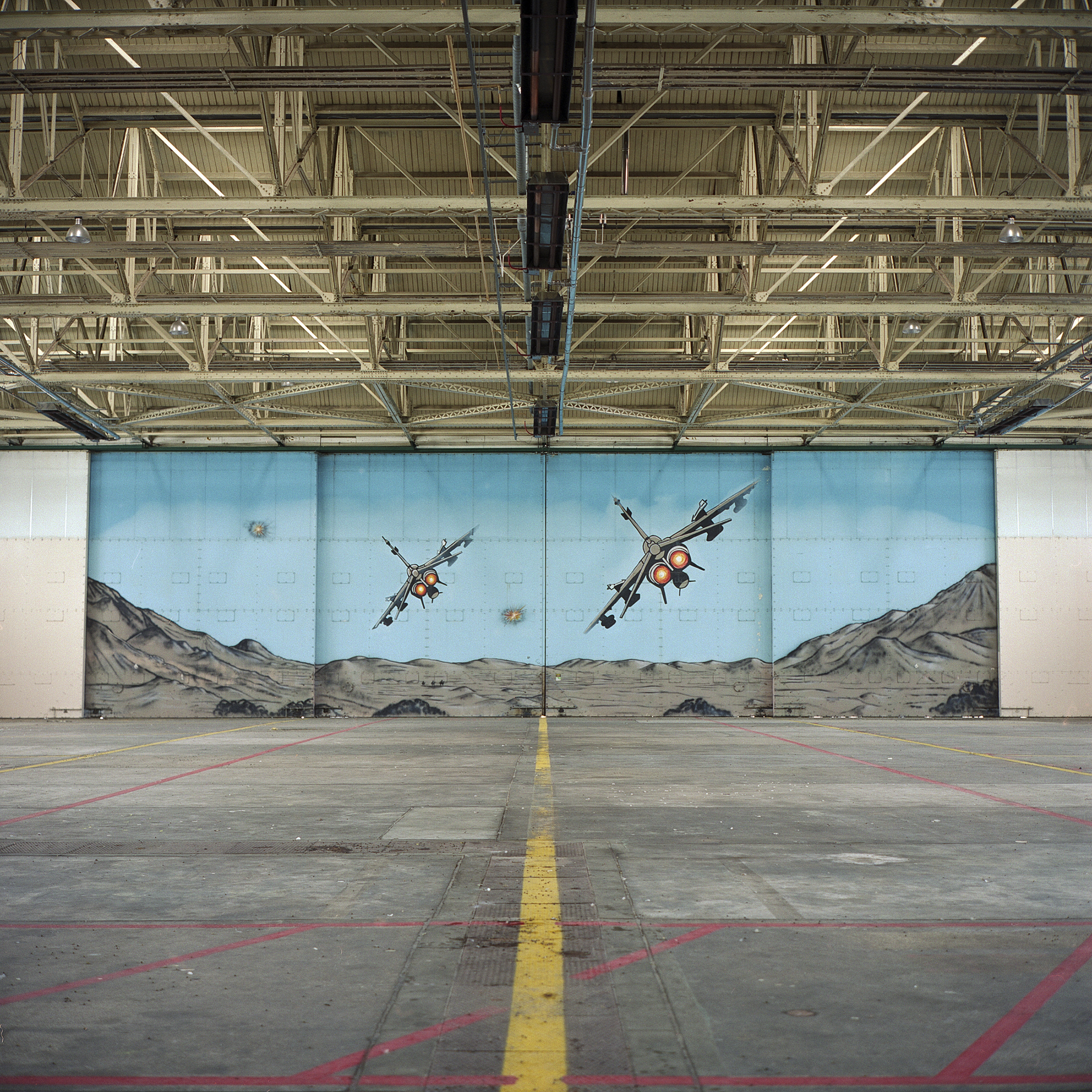 Yellow line (centered), with red lines that criss-cross, draws the eye to large hangar mural painted with two craft (in flight from behind) in a mountainous grey landscape