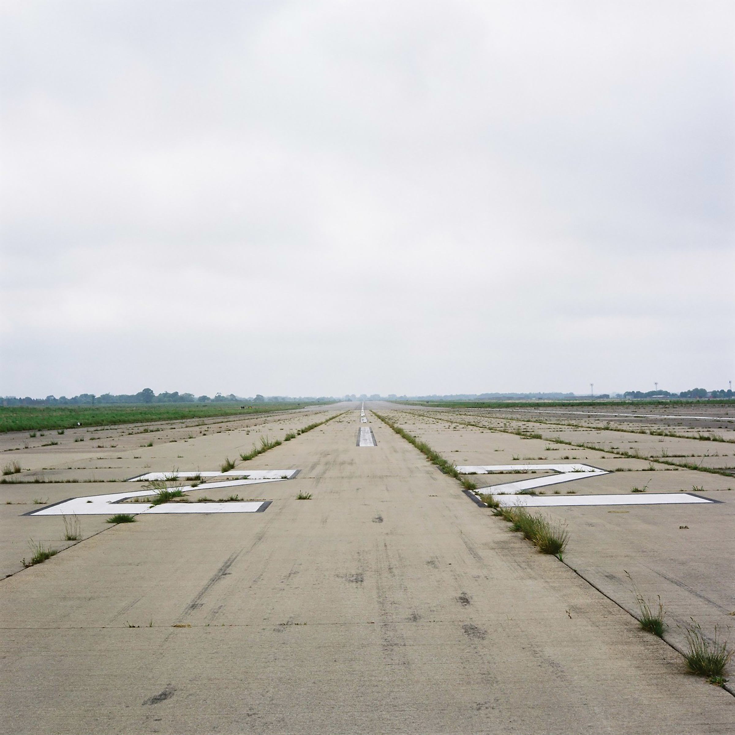 Grass grows between the concrete slabs on the runway which fades into vanishing point under a grey cloudy sky