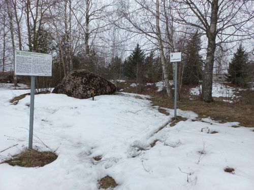 A large stone surrounded by snowy ground and trees, with two placards in the foreground