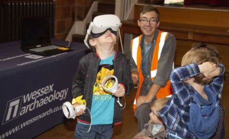 Two younger children and a smiling archaeologist look on as one child wearing the VR kit looks up while holdling the controllers