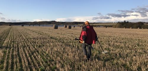 Photo of a person in a red jacket walks through a harvested field, holding a metal detector in their right hand and what appears to be a digging tool in their left. Round hay bales are scattered across the field, and in the background, a body of water is visible under a cloudy sky with patches of blue. Hills and trees line the distant horizon.